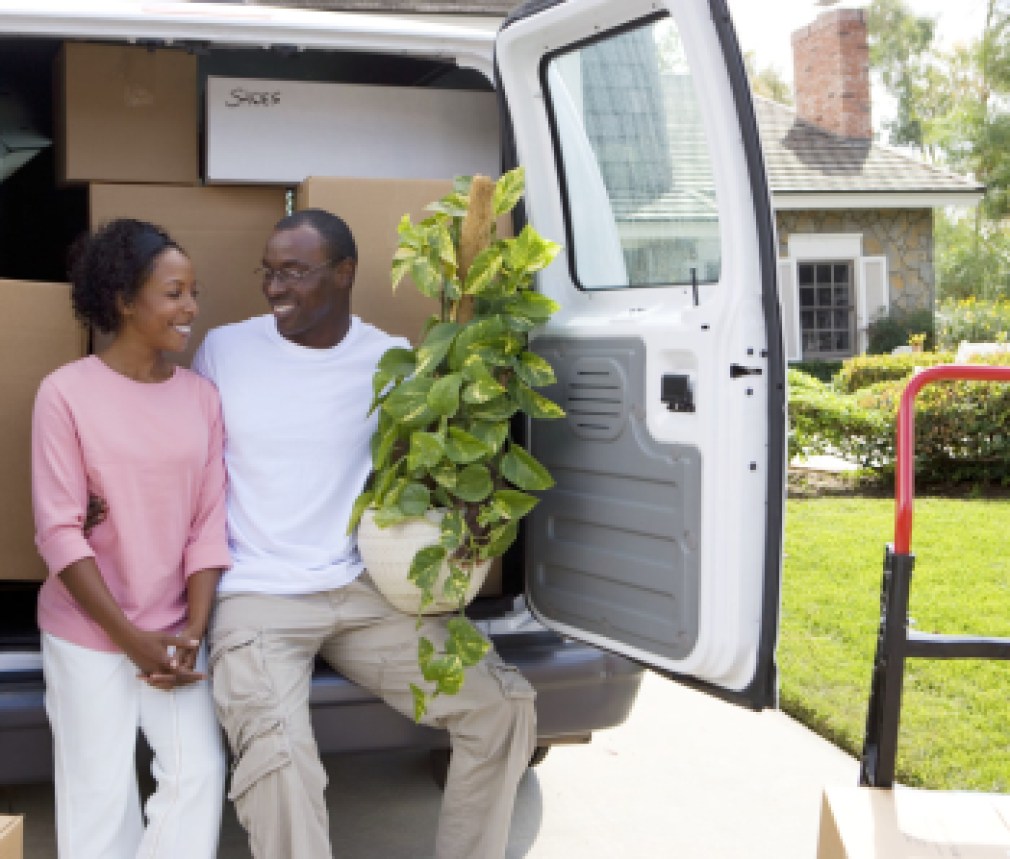 Smiling couple moving house in rear of van with pot plant at camera
