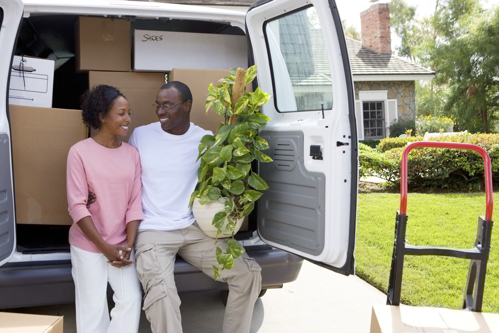 Smiling couple moving house in rear of van with pot plant at camera