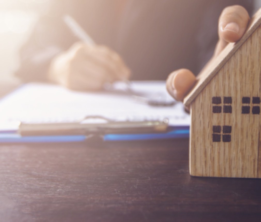 real estate, property and home owner signing contract concept, small wooden house model on office table with hand of woman buyer sign on rental agreement paper to rent above  mentioned residence