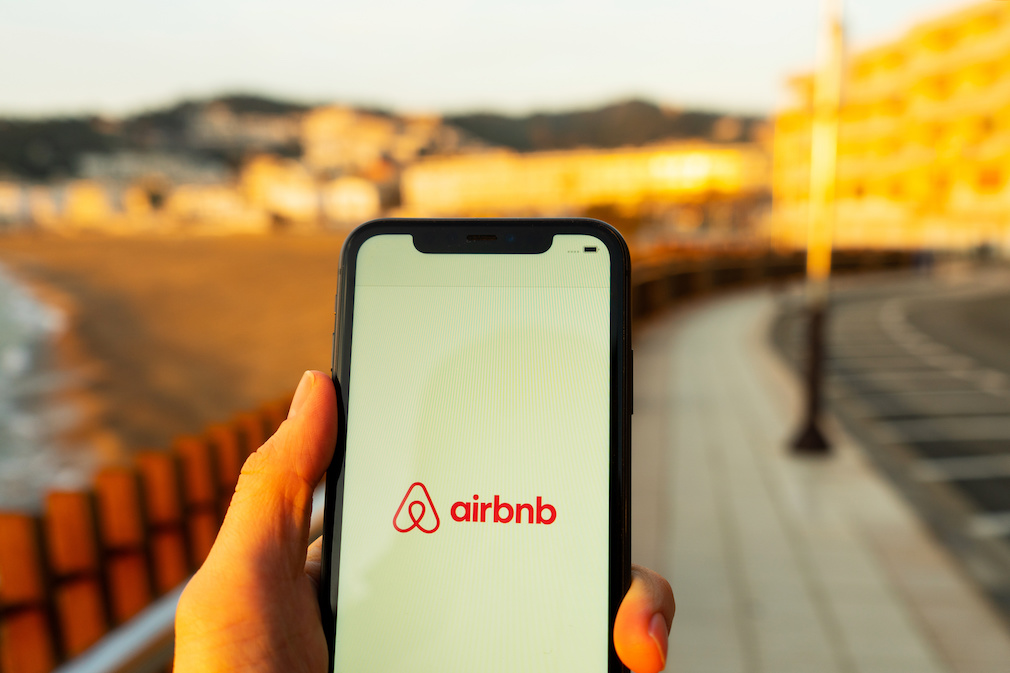 Hands holding iPhone 11 with an app of airbnb service. Holidays and vacation concept. A woman finding a place to stay or booking a hotel, apartment on a sunny day next to Mediterranean sea