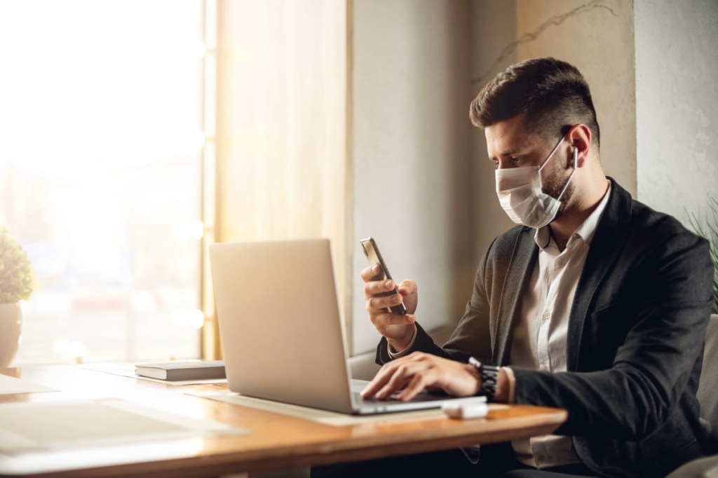 Caucasian adult bearded man indoors in cafe. Lifestyle concept photo with copy space. Picture with handsome guy who using protective face mask. Portrait with gray laptop