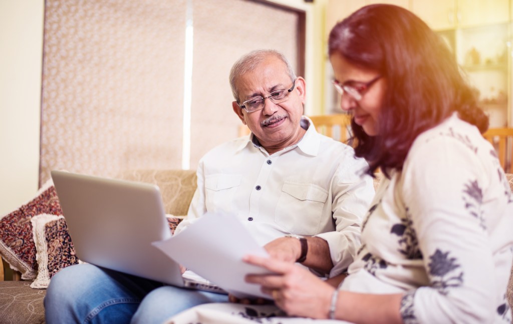 Senior Indian/asian couple accounting, doing home finance and checking bills with laptop, calculator and money while sitting on sofa/couch at home