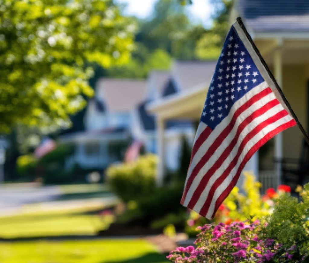 American flag waving in front of residential homes with green lawns and colorful flowers, symbolizing patriotism and community in suburban neighborhoods.